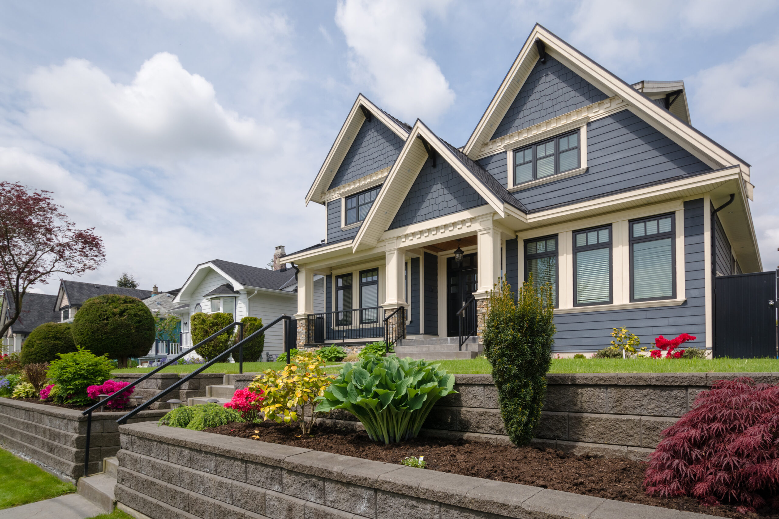 A lovely two-story house with gray-blue siding and an asphalt shingle roof with a garden in the front yard.