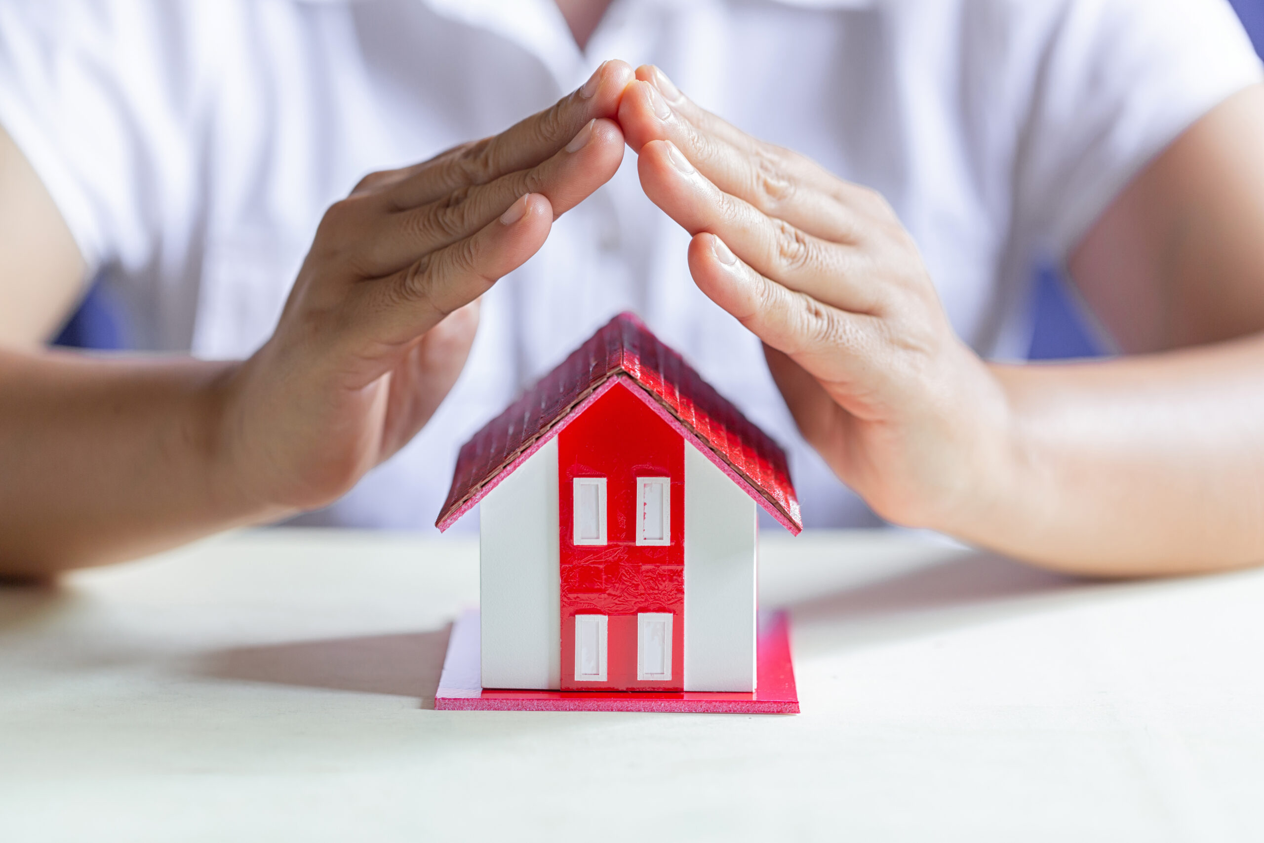 A small model of a white house with a red roof, with female hands tented over the top to signify insurance protection.