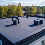 A Flat Roof With Ventilation Pipes And An Air Conditioning Unit Against A Forest Backdrop.